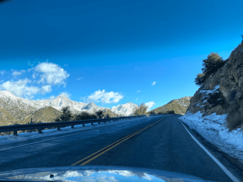 A scenic mountain road with snow on the ground, under a clear blue sky with a few clouds.