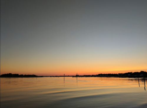 A serene sunset over calm waters, with silhouettes of distant land and poles reflecting in the water.