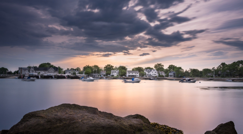A serene waterfront scene with boats on calm water, surrounded by trees and houses under a cloudy sunset sky.
