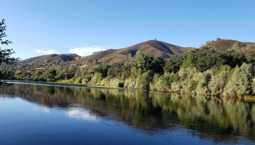 A serene river reflects lush greenery and rolling hills under a clear blue sky.