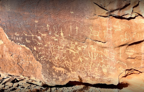 Ancient petroglyphs on a rock surface, depicting various figures, animals, and symbols in a desert landscape.