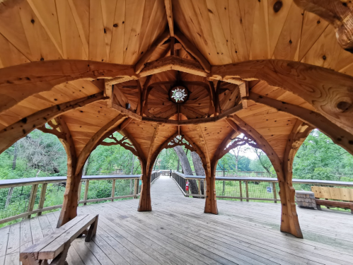 A wooden gazebo with intricate architecture, featuring a circular skylight and a view of surrounding greenery.