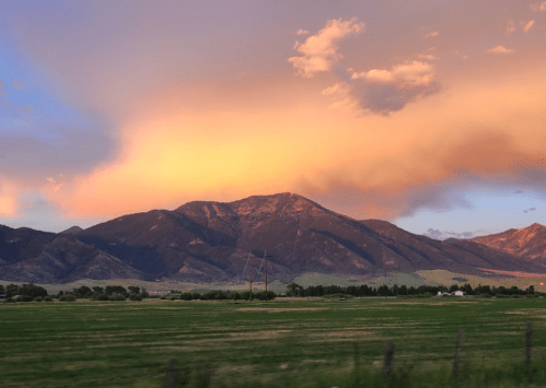 A scenic view of mountains under a colorful sunset, with green fields in the foreground and clouds in the sky.