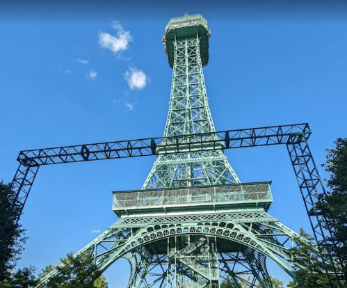 A tall green tower under a clear blue sky, framed by a metal structure, resembling the Eiffel Tower.