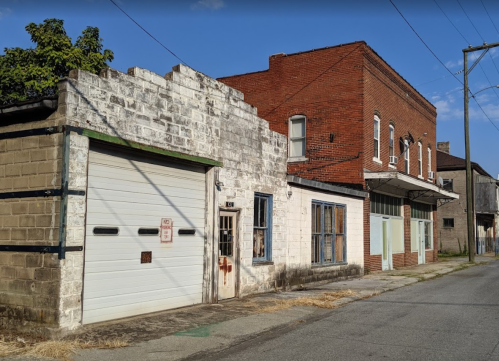 Abandoned buildings line a quiet street, featuring brick and weathered white walls under a clear blue sky.