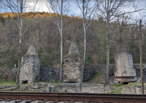 Three stone structures with conical tops, surrounded by trees and a hillside, near a railway track.