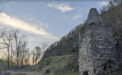 Ruins of a stone structure with a conical top, surrounded by trees and a sunset sky in the background.