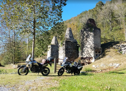 Two motorcycles parked near ancient stone ruins, surrounded by trees and greenery on a sunny day.