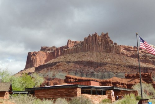 A red rock formation towers over a building, with an American flag waving in the foreground under a cloudy sky.