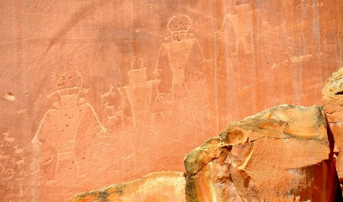Petroglyphs depicting human figures and symbols carved into a reddish rock surface.