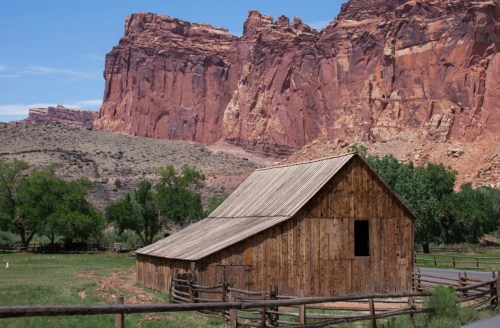A rustic wooden barn surrounded by green trees and towering red rock formations under a clear blue sky.