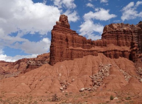 A towering red rock formation against a blue sky with scattered clouds, showcasing rugged desert terrain.