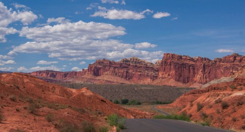 A winding road leads through red rock formations under a blue sky with fluffy clouds.