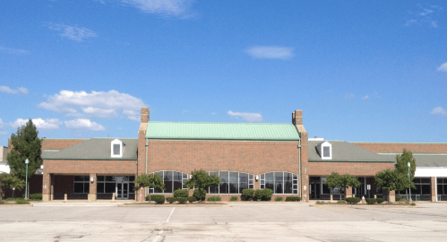 A brick building with a green roof, surrounded by trees, under a clear blue sky, in an empty parking lot.