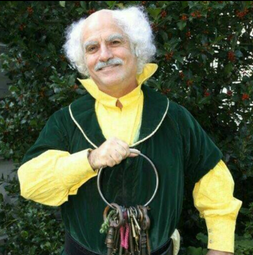 A man with curly white hair holds a large ring of keys, wearing a green velvet outfit and a yellow shirt, against a leafy background.