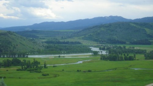 A scenic landscape featuring rolling green hills, a winding river, and distant mountains under a cloudy sky.