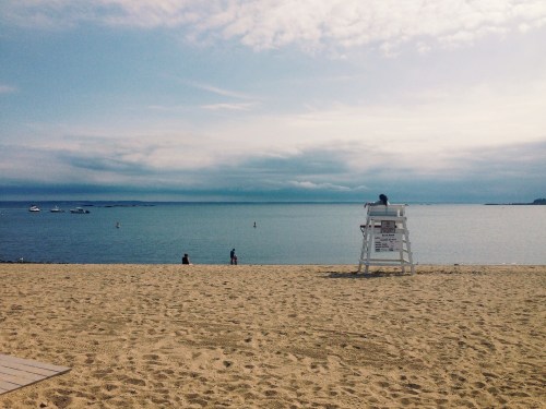 A lifeguard sits on a chair overlooking a calm beach with people and boats in the distance under a cloudy sky.