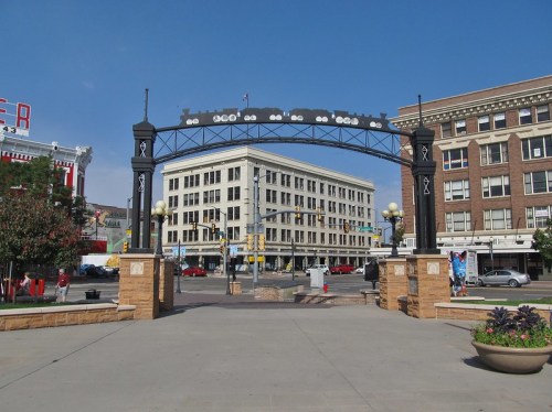 A decorative archway stands over a plaza with buildings and trees, under a clear blue sky.