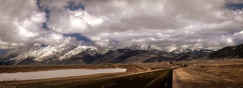 A panoramic view of snow-capped mountains under a cloudy sky, with a winding road in the foreground.