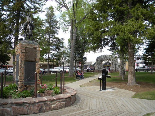 A park scene featuring a statue, trees, and a wooden walkway, with people sitting in the background.
