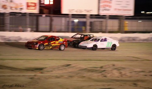 Three race cars compete on a dirt track at night, with colorful designs and bright lights in the background.