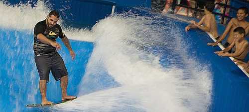 A surfer rides a wave on a flow rider while spectators cheer from the side.