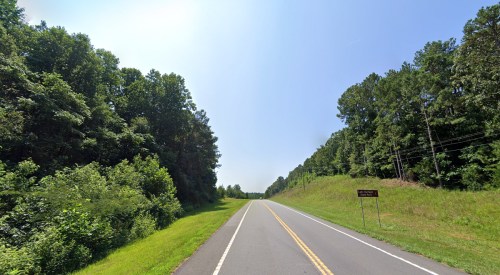 A straight road lined with trees on both sides under a clear blue sky. A sign is visible on the right.