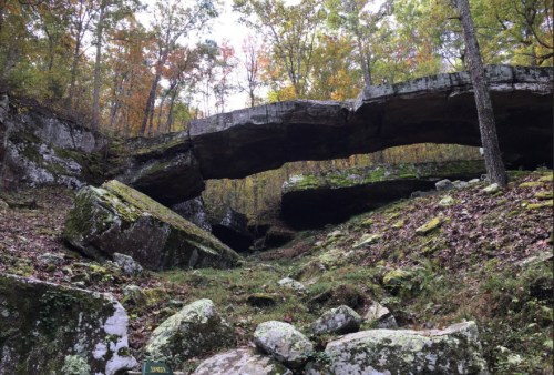 A natural rock arch surrounded by moss-covered boulders and trees in a forested area during autumn.