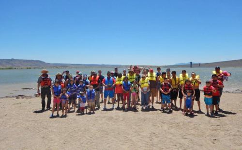 A large group of people in life jackets stands on a sandy beach by a lake on a sunny day.