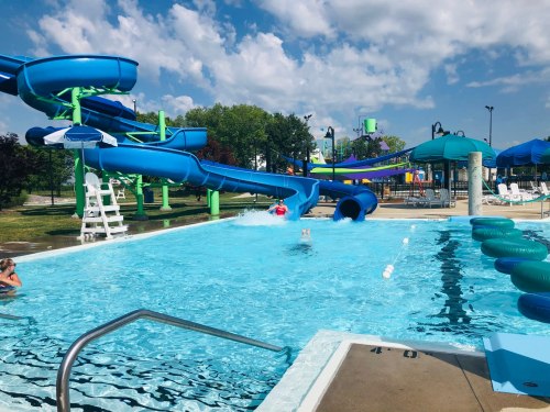 A water park scene featuring a blue slide, a swimming pool, and people enjoying the water on a sunny day.