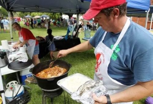 A man in a red cap stirs a large pot of food at an outdoor event, while another person prepares nearby.