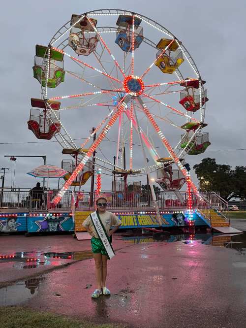 A girl in a sash stands in front of a brightly lit Ferris wheel at a fair, with a cloudy sky and wet ground.