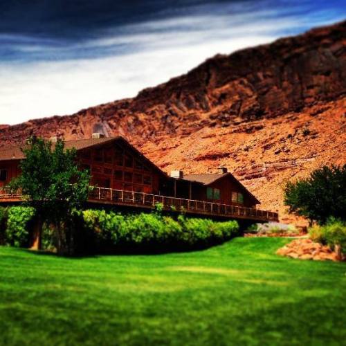 A rustic lodge surrounded by lush greenery and rocky mountains under a blue sky with wispy clouds.