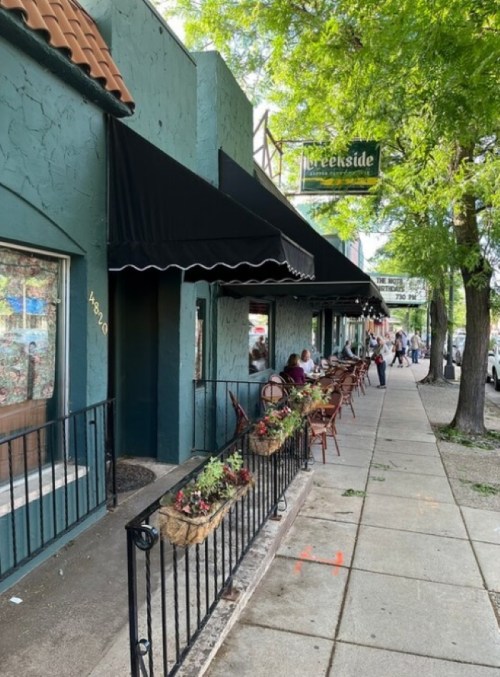A green storefront with black awnings, outdoor seating, and planters along a sidewalk lined with trees.