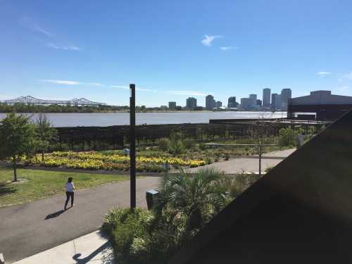 A person walks along a path with a view of a river and city skyline under a clear blue sky.