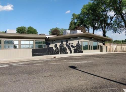 A building with a mural depicting figures engaged in various activities, surrounded by trees and a clear blue sky.