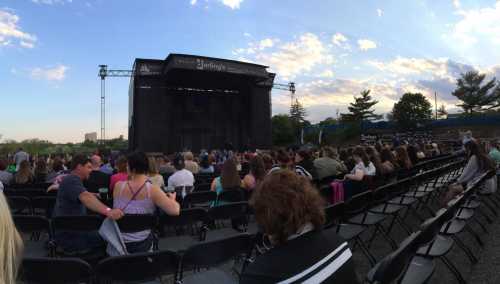 A crowd of people sits in front of a large, empty stage under a blue sky with scattered clouds.