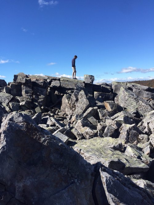 A person stands on a rocky outcrop under a clear blue sky, surrounded by large stones and boulders.