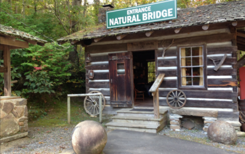 A rustic log cabin with a sign reading "Entrance Natural Bridge," surrounded by greenery and stone features.