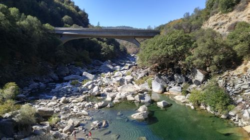 A bridge spans a rocky riverbed surrounded by lush greenery under a clear blue sky.