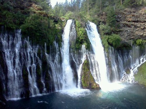 A scenic view of two waterfalls cascading into a clear pool, surrounded by lush greenery and rocky cliffs.