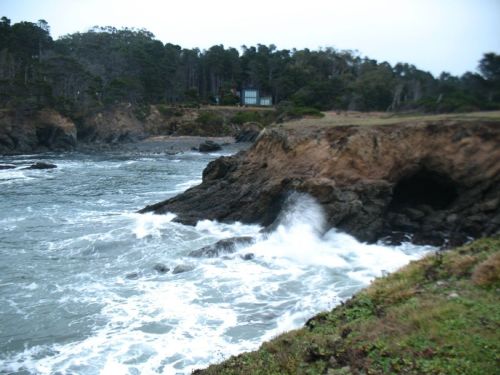 Waves crash against rocky cliffs along a coastal shoreline, with trees and a house visible in the background.