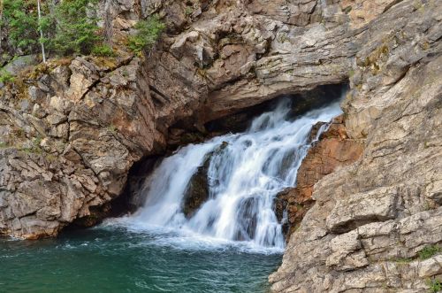 A serene waterfall cascading over rocky cliffs into a tranquil green pool below, surrounded by lush greenery.