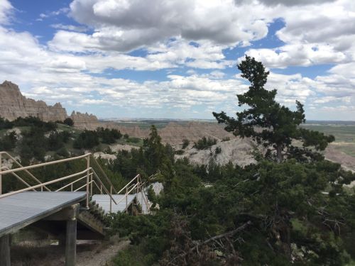A scenic view of rugged cliffs and valleys under a cloudy sky, with a wooden walkway and trees in the foreground.