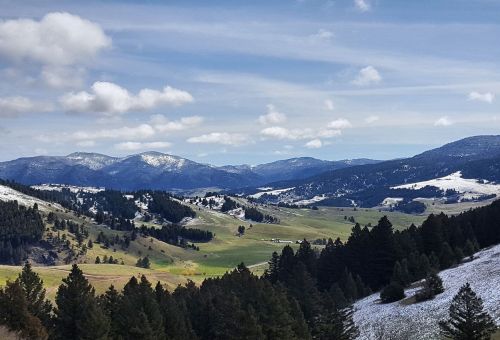 A scenic view of rolling green hills and mountains under a partly cloudy sky, with patches of snow on the peaks.