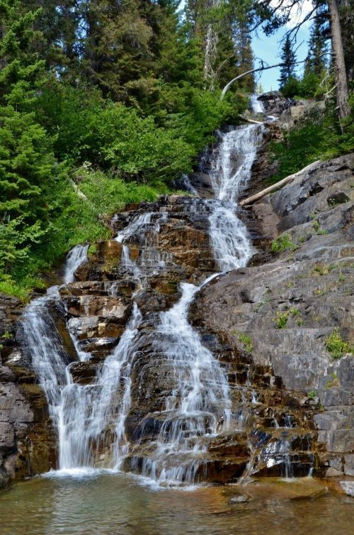 A serene waterfall cascading down rocky terrain, surrounded by lush green trees and a clear blue sky.