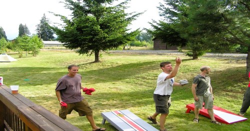 Three men play a lawn game in a grassy area surrounded by trees, with a tent and buildings in the background.