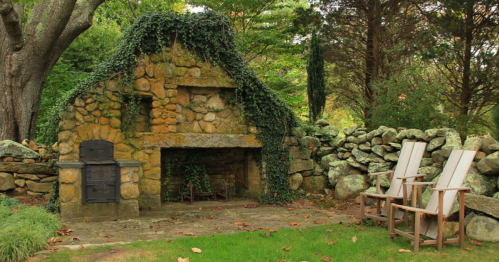 A rustic stone fireplace covered in ivy, surrounded by a stone wall and two wooden chairs in a serene garden setting.