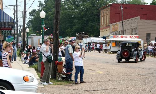 A vintage car drives by as a crowd of people, some with cameras, watch from the street during a local event.