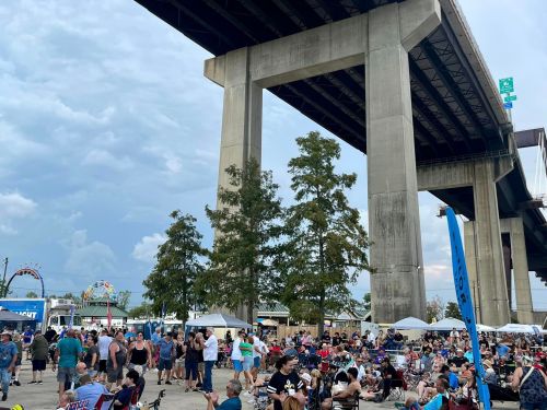 A crowded outdoor event under a large bridge, with people gathered around food stalls and a carnival in the background.
